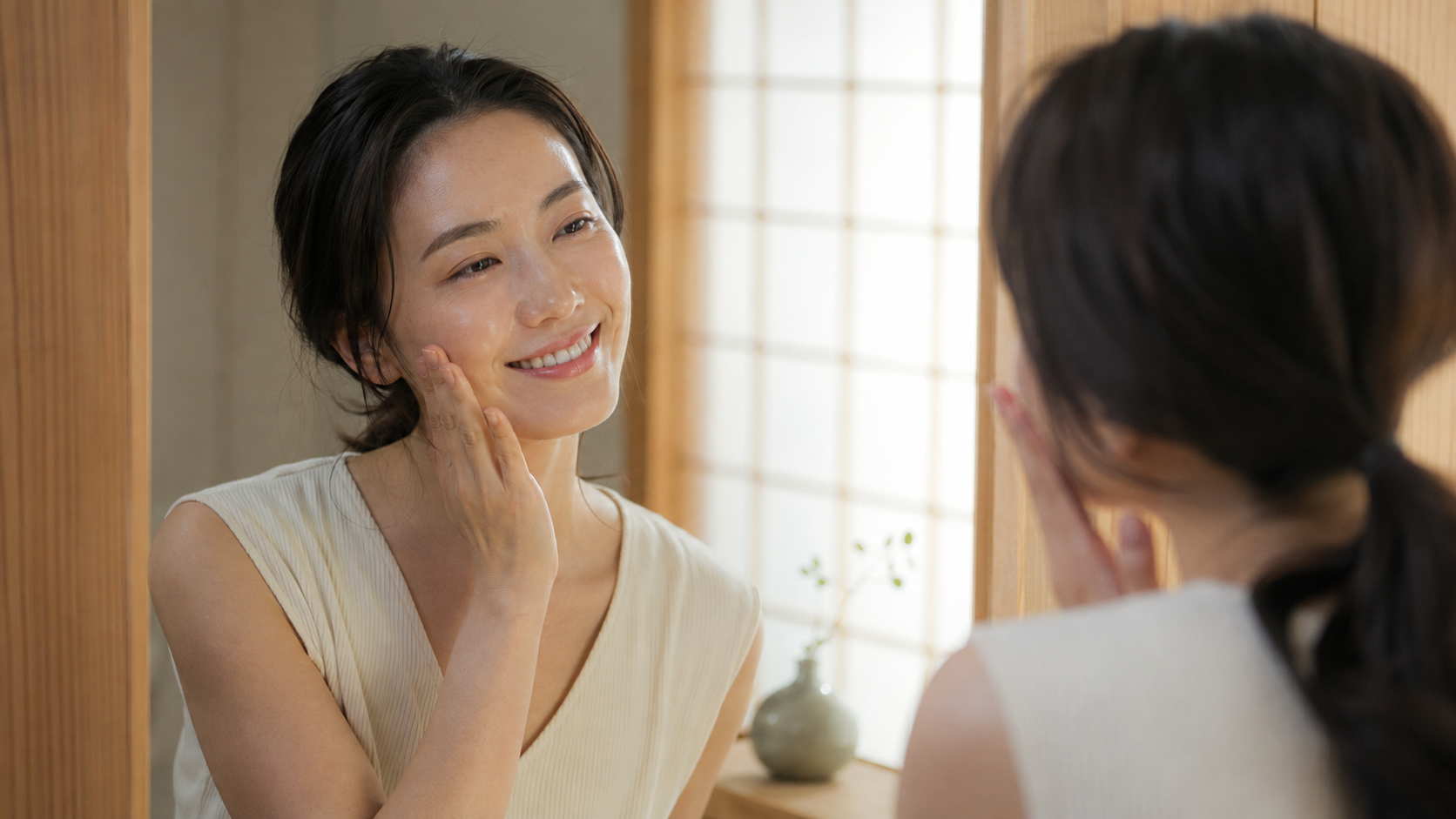 Japanese woman smiling softly while pressing hydration into her cheeks in front of a mirror.